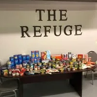 the-refuge-logo A table of packaged foods sitting on in front of a wall with the food pantry name of "The Refuge".