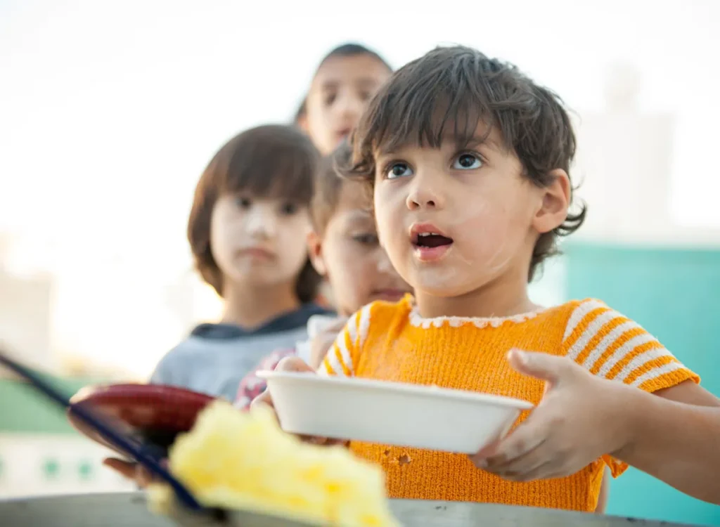 Photo of young children, about 5 to 7 years old, waiting in line to get food, holding a bowl.