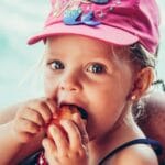This is Who We Are: Close-up of a cute Caucasian child enjoying fruit while wearing a vibrant pink cap with a mermaid design.