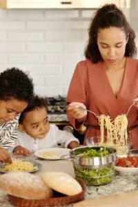 A mom dishing up pasta for her two children at the dinner table with bread and vegetables close by.