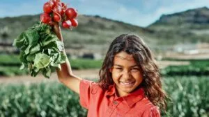 Smiling girl holding red radishes in a field under a blue sky during daytime.