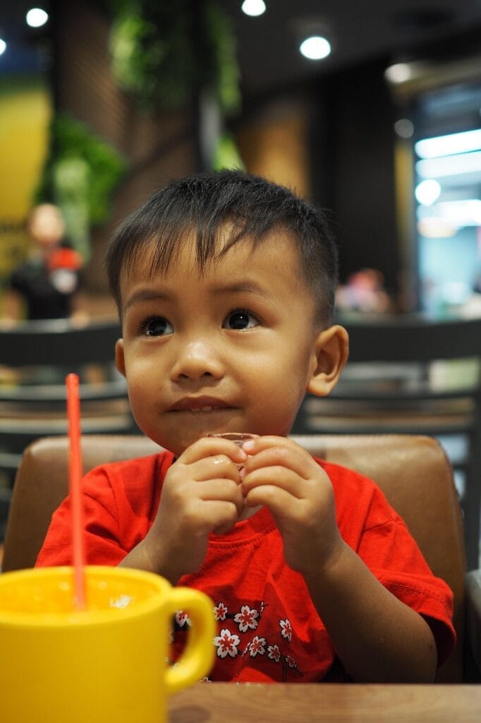 Young male toddler of Asian descent sitting at a restaurant eating a cracker with a yellow cup of juice in front of him