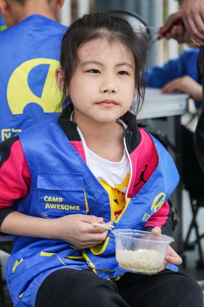 Young female asian child at camp with a blue vest on sitting and eating a bowl of rice.